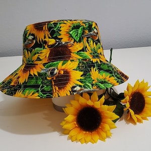 May include: A bucket hat with a vibrant sunflower and bird pattern. The hat features large yellow sunflowers with brown centers, green leaves, and small birds against a black background. The hat is displayed with two artificial sunflowers.