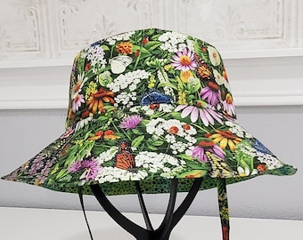 May include: A floral print bucket hat with a colourful pattern of butterflies, flowers, and foliage in green, orange, purple, and white. The hat has a chin strap with a black toggle. It is displayed on a black stand.