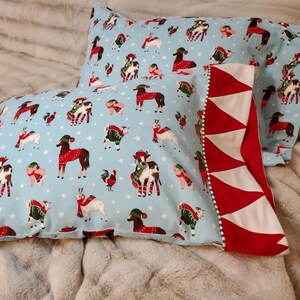 May include: Two light blue pillowcases with a festive pattern of animals wearing scarves and hats. The pillowcases have a red and white geometric border. The background is a soft, neutral color.