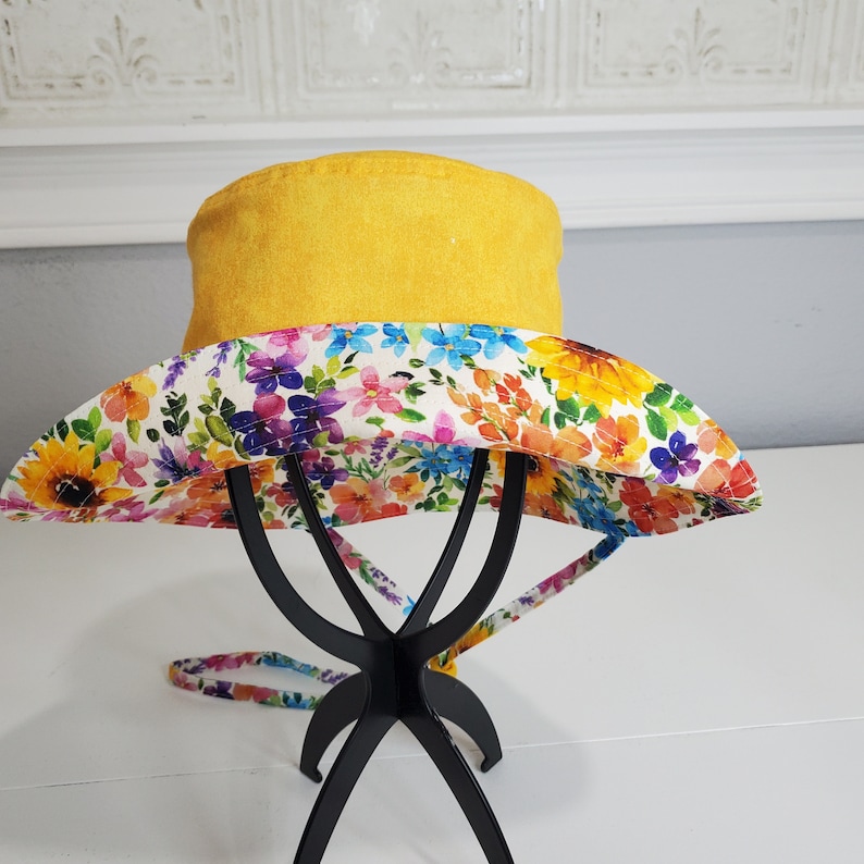 May include: A yellow bucket hat with a floral print brim. The brim features a colourful pattern of flowers in shades of pink, blue, yellow, and orange. The hat is displayed on a black metal stand.