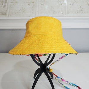 May include: A yellow bucket hat with a floral patterned brim and matching chin strap. The hat is displayed on a black stand against a white background. The chin strap has a yellow bead.