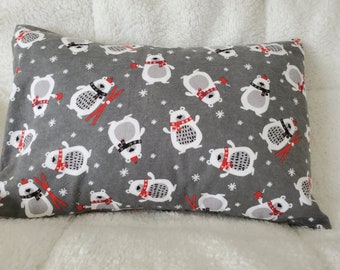 May include: Gray rectangular pillow with a repeating pattern of white polar bears wearing red scarves and hats, some with skis. The bears are interspersed with white snowflakes. The pillow is on a white, fluffy surface.