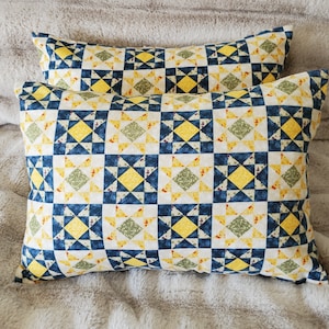 May include: Two rectangular throw pillows with a patchwork design. The pattern features squares with yellow, blue, and green star and diamond shapes on a white background. The pillows are resting on a gray, textured surface.