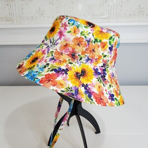 May include: A floral bucket hat with a bright watercolour design. The hat displays yellow sunflowers, orange, pink, and purple flowers on a white background. It has a wide brim and matching ties.