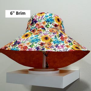 May include: A wide-brimmed sun hat with a floral pattern featuring sunflowers and other colorful blooms. The brim measures 15 cm. The underside of the brim is a solid orange color.