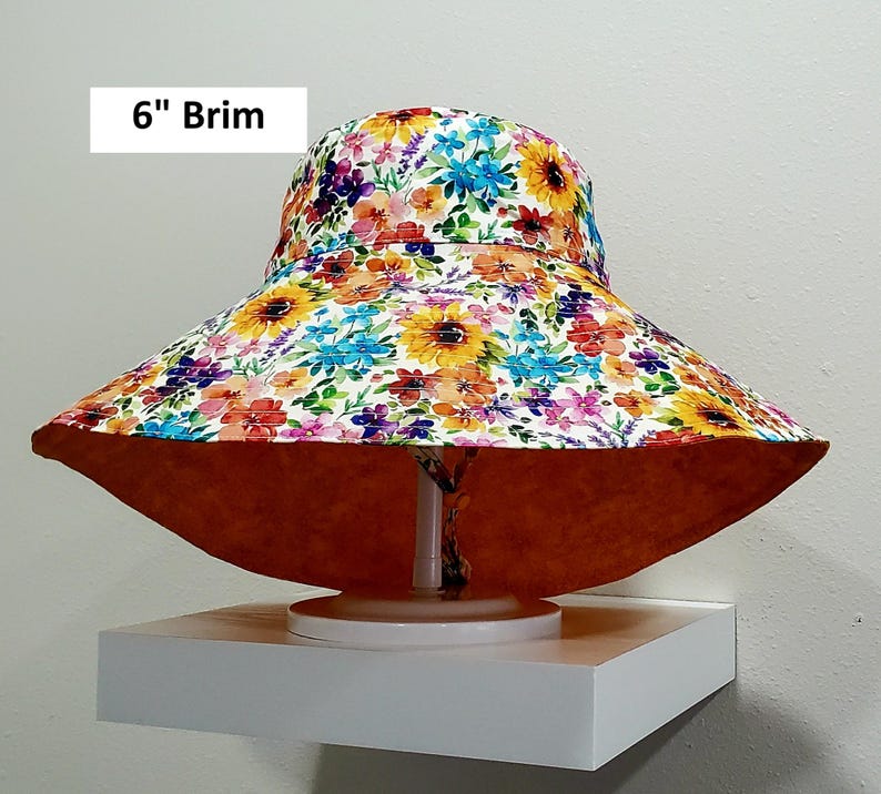 May include: A wide-brimmed sun hat with a floral pattern featuring sunflowers and other colorful blooms. The brim measures 15 cm. The underside of the brim is a solid orange color.