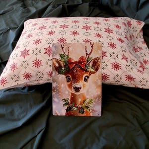 May include: A rectangular metal sign featuring a brown deer with large eyes, a red bow, and holly leaves on its head, set against a snowy background. Behind the sign is a white pillowcase with a repeating pattern of red snowflakes and green sprigs.