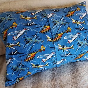 May include: Rectangular pillow with a blue background covered in illustrations of various aircraft, including airplanes and helicopters. The pillow is made of fabric and has a seam down the middle.