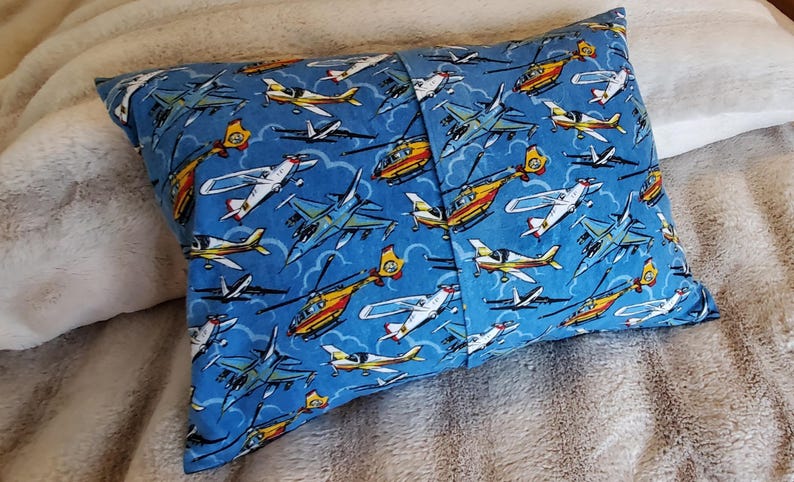 May include: Rectangular pillow with a blue background covered in illustrations of various aircraft, including airplanes and helicopters. The pillow is made of fabric and has a seam down the middle.