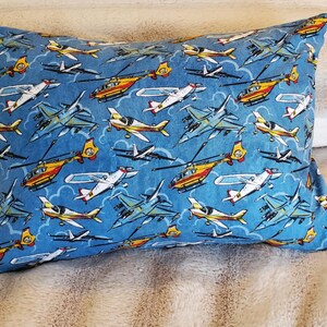 May include: A rectangular pillow with a blue background featuring a pattern of various airplanes and helicopters in shades of yellow, white, and gray. The pillow is resting on a soft, textured surface.