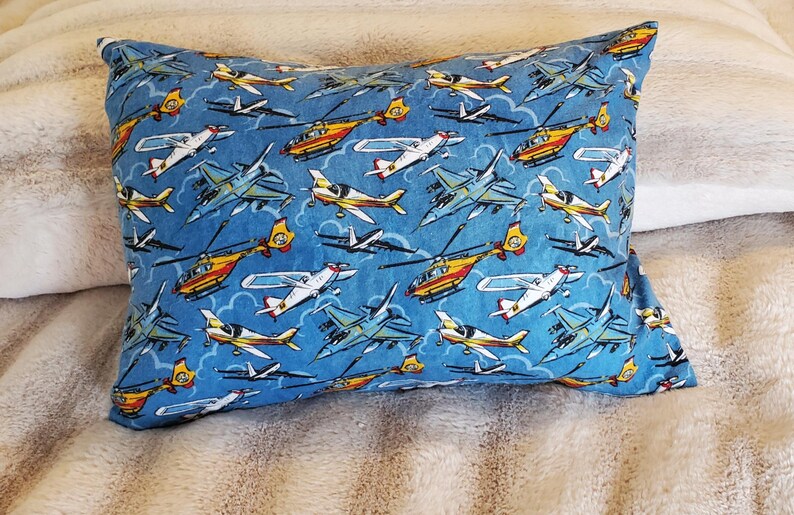 May include: A rectangular pillow with a blue background featuring a pattern of various airplanes and helicopters in shades of yellow, white, and gray. The pillow is resting on a soft, textured surface.