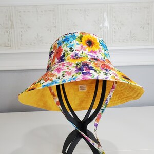 May include: A reversible bucket hat with a vibrant floral pattern featuring sunflowers, blue, red, and purple flowers on a white background. The interior is a solid yellow, and it has matching floral straps. The hat is displayed on a black stand.