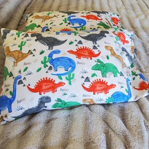 May include: Two white pillowcases with a colorful dinosaur pattern. The design includes various dinosaurs in blue, red, green, brown, and gray, along with cacti and the word "Roar" in black. The pillowcases are on a soft, textured surface.