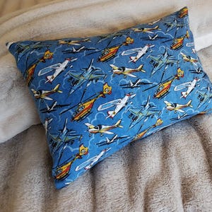 May include: A rectangular pillow with a blue background and a pattern of various aircraft, including airplanes and helicopters. The aircraft are in shades of yellow, white, and blue. The pillow is resting on a soft, light-colored blanket.