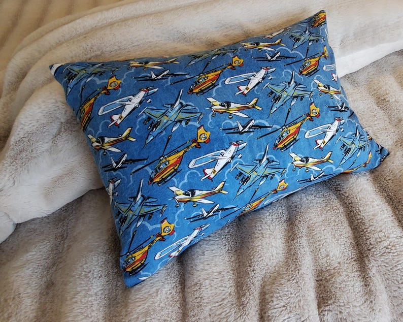 May include: A rectangular pillow with a blue background and a pattern of various aircraft, including airplanes and helicopters. The aircraft are in shades of yellow, white, and blue. The pillow is resting on a soft, light-colored blanket.