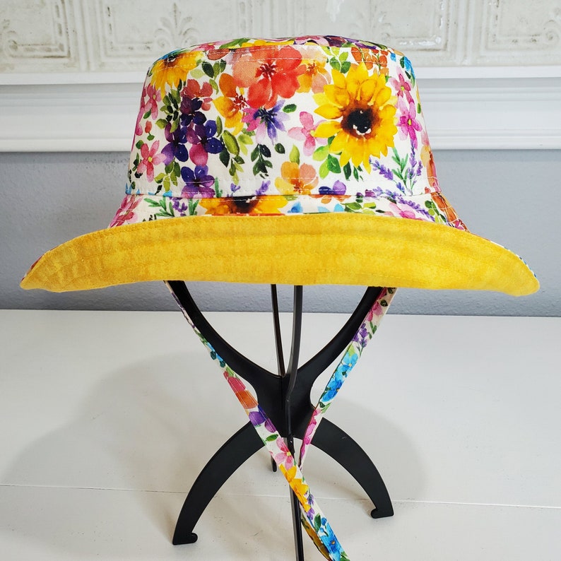 May include: A floral bucket hat with a bright yellow brim and a colourful floral pattern. The hat features a mix of red, yellow, purple, and green flowers on a white background. The hat is displayed on a black stand with floral patterned straps.
