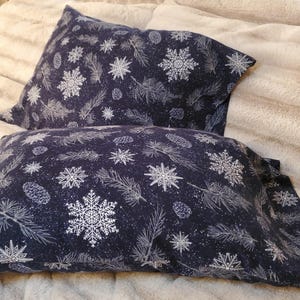May include: Two navy blue pillowcases with a white snowflake and pine needle pattern. The pillowcases are rectangular and rest on a beige, textured surface. The design includes snowflakes, pine needles, and pine cones.