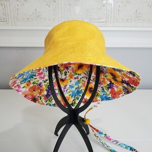 May include: A reversible sun hat. The exterior is bright yellow, and the interior is patterned with colorful flowers. It has a wide brim and a chin strap with a yellow bead. The hat is designed for sun protection.