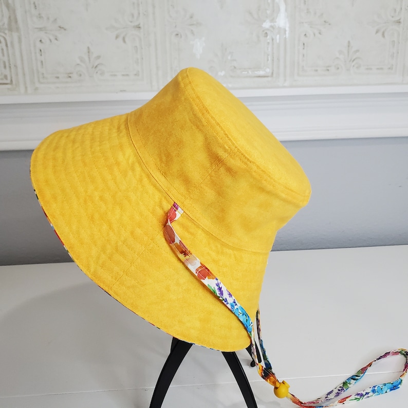 May include: A yellow bucket hat with a floral lining and a matching chin strap. The hat is made of a textured fabric and has a wide brim. The chin strap is adjustable with a yellow bead.