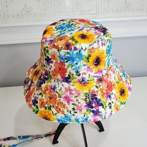May include: A floral bucket hat with a bright pattern of sunflowers and other colourful flowers in yellow, orange, red, blue, and purple. The hat has a wide brim and a matching fabric tie. The hat is displayed on a black stand.