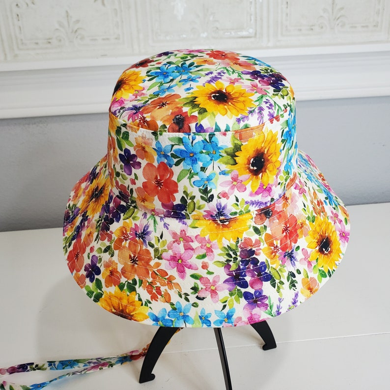 May include: A floral bucket hat with a bright pattern of sunflowers and other colourful flowers in yellow, orange, red, blue, and purple. The hat has a wide brim and a matching fabric tie. The hat is displayed on a black stand.
