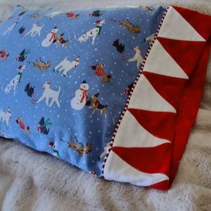 May include: A rectangular pillow with a light blue background featuring a pattern of dogs and snowmen. The pillow has a red and white striped border on one side. The pillow is resting on a soft, white blanket.