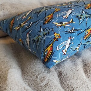 May include: A rectangular pillow with a blue background and a pattern of various airplanes and helicopters in white, yellow, and orange. The pillow is resting on a soft, gray, textured surface.