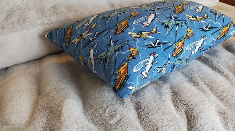 May include: A rectangular pillow with a blue background and a pattern of various airplanes and helicopters in white, yellow, and orange. The pillow is resting on a soft, gray, textured surface.