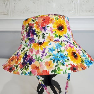 May include: A floral bucket hat with a bright pattern of sunflowers, lavender, and other colourful flowers. The hat has a wide brim and matching ties. The background is a white surface.