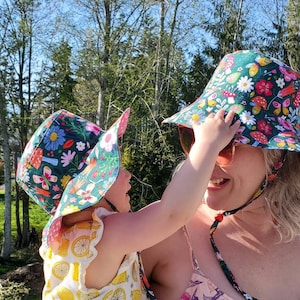 May include: Two floral print sun hats, one on a child and one on an adult. The hats feature a dark green background with colorful flowers, mushrooms, and butterflies. The child is wearing a yellow dress with lemon graphics.