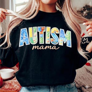 May include: Black sweatshirt featuring the word "AUTISM" in patchwork letters of various colours, and the word "mama" in cursive. The letters are blue, yellow, and green. The sweatshirt is a crew neck style.