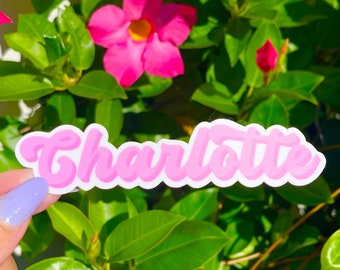 May include: A die-cut vinyl sticker featuring the name Charlotte written in a bubble-style, light pink font with a white outline. The sticker is held against a background of vibrant green leaves and bright pink mandevilla flowers.