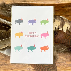 May include: A white card with a birthday message that says "Herd it's your birthday!" and features six illustrations of buffalo wearing party hats in different colors.