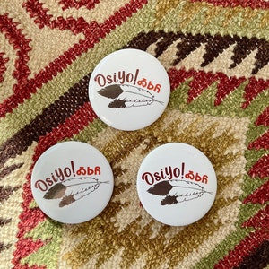 May include: Three white round buttons with a red and black feather design and the text "Osiyo!" in red. The buttons are on a brown and red patterned fabric.