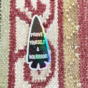 May include: A black and silver holographic sticker shaped like an arrowhead with the text "Prove Yourself A Warrior".