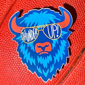 May include: A die-cut sticker featuring a blue bison wearing sunglasses with the words "THUNDER UP!" in white. The bison has orange horns and a red nose. The sticker is on a red textured surface.