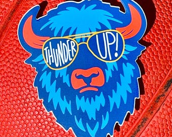 May include: A die-cut sticker featuring a blue bison wearing sunglasses with the words "THUNDER UP!" in white. The bison has orange horns and a red nose. The sticker is on a red textured surface.