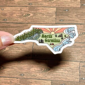 North Carolina sticker featuring Charlotte, Raleigh/Durham area, mountains, beaches, sunshine, and farms. Sticker is in the shape of NC with a white offset.