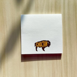 May include: A square white sticky note featuring a dark brown silhouette of a bison. Inside the bison shape, the word CHOCTAW is printed in bold yellow capital letters. The paper has a smooth texture and a clean, straight-edged square format.