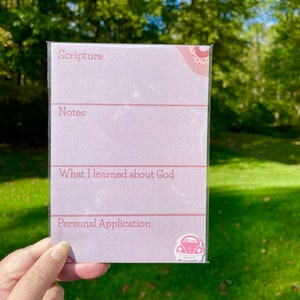 May include: A pink and white notepad with the words "Scripture," "Notes," "What I learned about God," and "Personal Application." The notepad has a decorative design in the upper right corner and a small logo in the bottom right corner.