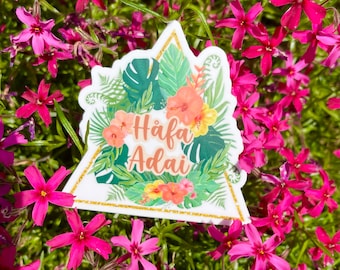 May include: A white triangle sticker with a gold glitter border and tropical floral design. The sticker says "Häfa Adai" in gold lettering.