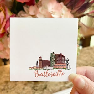 May include: A white card with a hand-drawn illustration of the Bartlesville, Oklahoma skyline. The city name is written in pink below the illustration.