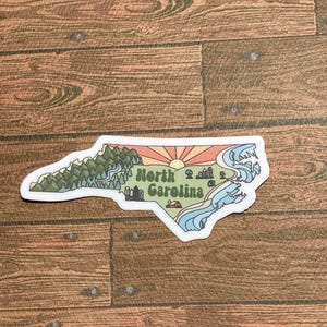 North Carolina sticker featuring Charlotte, Raleigh/Durham area, mountains, beaches, sunshine, and farms. Sticker is in the shape of NC with a white offset.