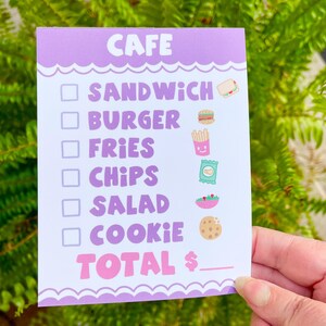May include: A purple and white cafe menu with a checklist of food items including sandwich, burger, fries, chips, salad, and cookie. The menu has a pink "TOTAL $" with a line for the total amount.