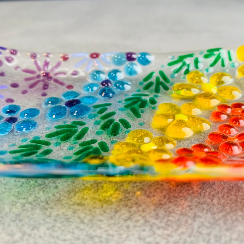 Puede incluir: Un plato de vidrio transparente pintado con un arco&iacute;ris de colores. El plato est&aacute; decorado con flores y hojas.