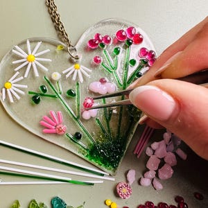 May include: A heart-shaped glass pendant with a floral design, featuring pink, red, and white flowers, green stems, and yellow centers. A gold chain is attached. The pendant is being assembled with tweezers, surrounded by glass pieces.