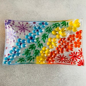 Puede incluir: Un plato rectangular de vidrio con un dise&ntilde;o floral arco&iacute;ris. El plato est&aacute; decorado con peque&ntilde;os trozos de vidrio en tonos de morado, azul, verde, amarillo, naranja y rojo, formando un patr&oacute;n de flores y hojas.