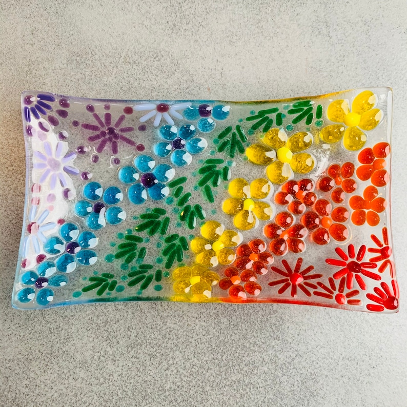 Puede incluir: Un plato rectangular de vidrio con un dise&ntilde;o floral arco&iacute;ris. El plato est&aacute; decorado con peque&ntilde;os trozos de vidrio en tonos de morado, azul, verde, amarillo, naranja y rojo, formando un patr&oacute;n de flores y hojas.