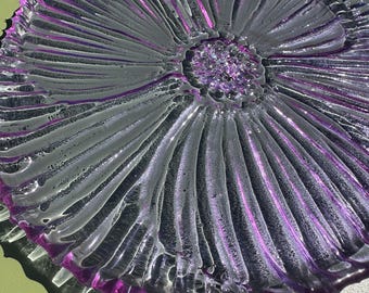 May include: A decorative, clear glass plate with a flower-like design. The plate features a central, textured area surrounded by radiating ridges, with a vibrant purple hue accenting the design. The plate's edges are also outlined in purple.