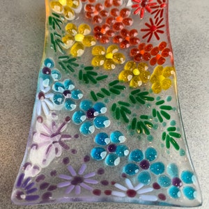 Puede incluir: Un plato de vidrio transparente con un dise&ntilde;o floral de arco&iacute;ris. El plato est&aacute; decorado con peque&ntilde;as flores y hojas de vidrio de colores.